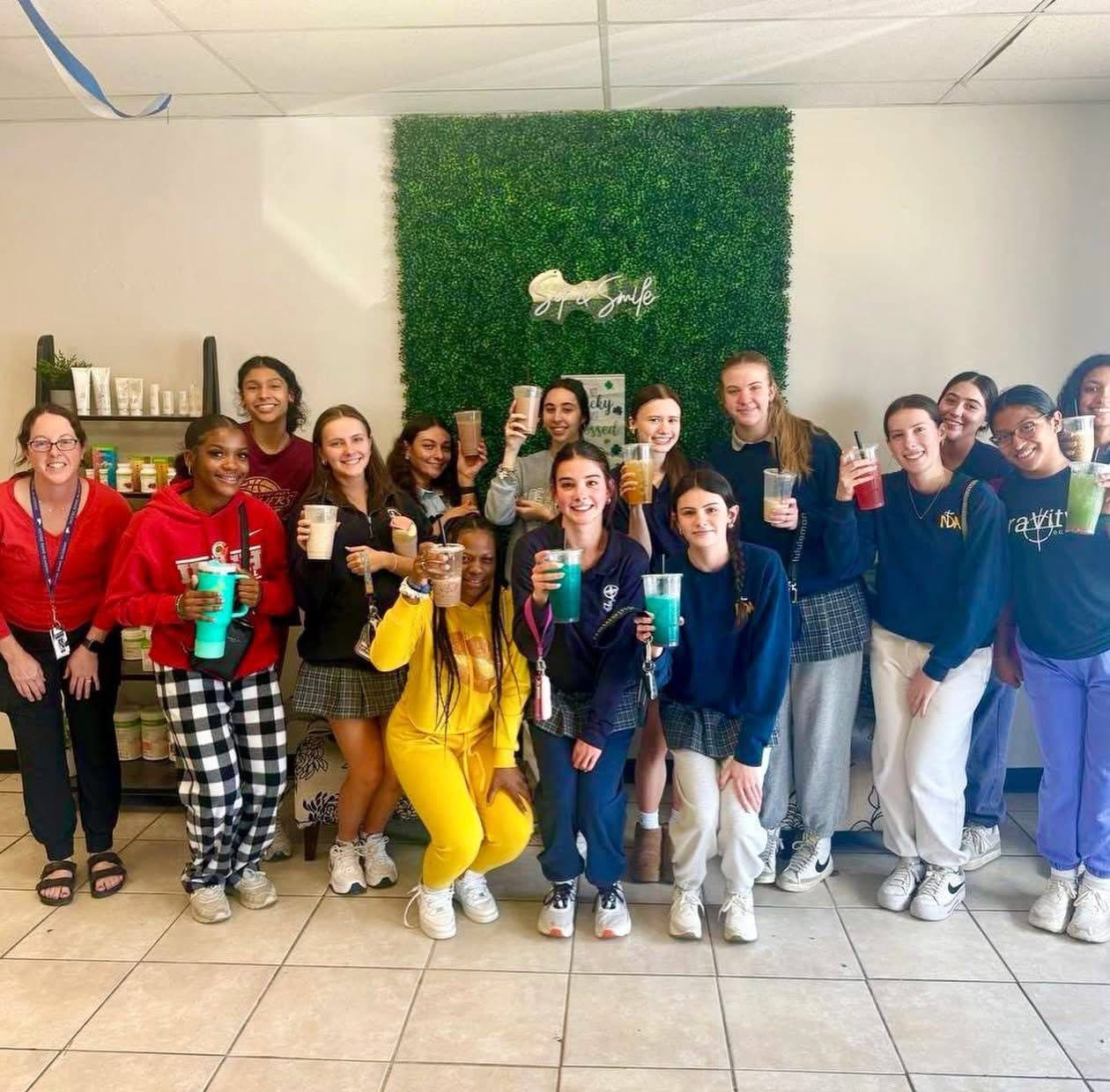 A big group of smiling regulars raising their shakes and teas in front of NutriToledo's Sip & Smile wall